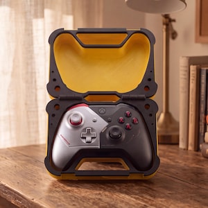 Può includere: Una custodia per controller di videogiochi gialla e nera, con design a cerniera. All'interno è visibile un controller wireless bicolore argento e nero, con accenti rossi sui pulsanti e sul joystick e un piccolo logo rosso sul lato sinistro.