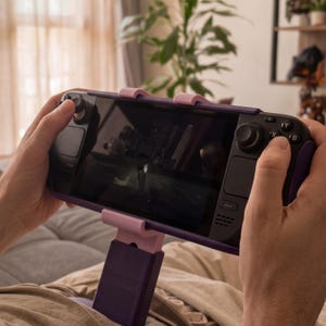 以下が含まれることがあります： 黒い画面とダークパープルの筐体を持つ携帯型ゲーム機。パープルとライトピンクの調節可能なスタンドに固定されています。2つのアナログスティック、方向パッド、A、B、X、Yとラベル付けされた4つのアクションボタンを備えています。