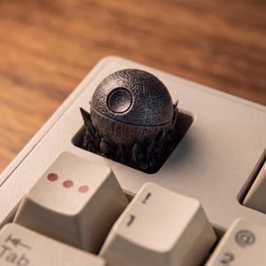 Può includere: Un tasto meccanico personalizzato raffigurante un dettagliato modello di stazione spaziale sferica grigio metallizzato con una cupola concava, montato su una base cristallina scura e frastagliata su una tastiera beige.