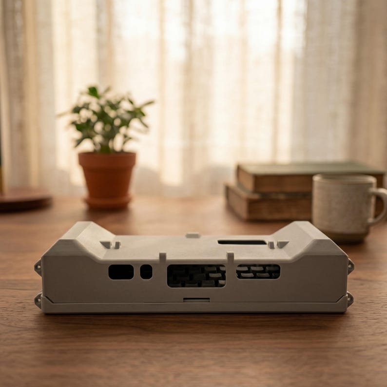 May include: A light gray rectangular plastic enclosure featuring a series of cutouts and ports along its front edge, including a large central opening with internal grid-like structures and smaller circular and rectangular ports on the sides.