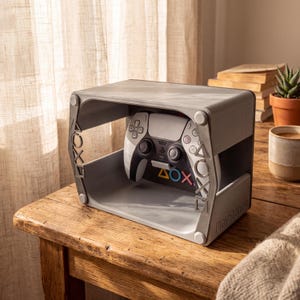 May include: A gray, rectangular display stand for a video game controller, featuring the PlayStation logo and iconic triangle, circle, cross, and square symbols on the side panels and back. A white and black game controller rests inside.