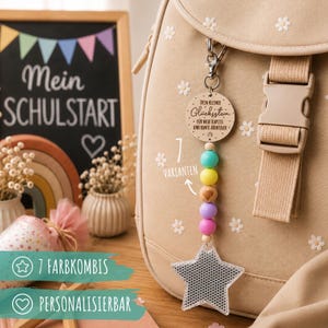 Könnte beinhalten: Ein beiger Rucksack mit weißen Blumenmustern ist mit einem hängenden Schlüsselanhänger versehen. Dieser besteht aus einer runden Holzscheibe mit deutschem Text, einer Reihe bunter Perlen in Türkis, Gelb, Naturholz, Lavendel und Pink sowie einem sternförmigen Reflektor.