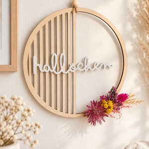 Könnte beinhalten: Ein kreisförmiger Wandbehang aus Holz mit vertikalen Lamellen auf der linken Hälfte und dem weißen Schriftzug "hallöchen" in der Mitte. Die rechte Seite ist mit einem Bündel getrockneter Blumen in Rosa-, Gelb- und Violetttönen verziert.