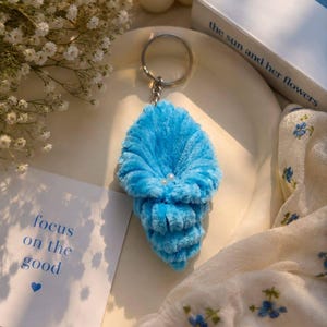 May include: A bright blue plush keychain shaped like a seashell with a soft, ribbed texture. The center of the shell features two small, round white pearls. The keychain is attached to a standard silver-toned metal split ring and a short chain.