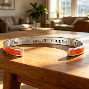 May include: A silver-toned cuff bracelet featuring a vibrant orange enamel inlay along the outer band. The interior surface is engraved with the black text, "This too shall pass, BUT FUCKING HELL," in a clear, serif font style.