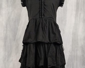 May include: A black knee-length dress featuring a high ruffled collar, short cap sleeves with ruffled edges, and a button-down front placket. The skirt is constructed with three horizontal tiers of fabric, creating a layered, voluminous silhouette.