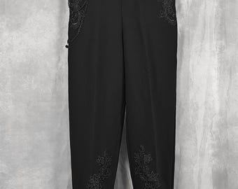 May include: Black trousers featuring intricate black floral lace embroidery on the upper hip and lower leg areas. The pants include a decorative black metal chain attached to the right hip pocket area and a subtle lace trim at the bottom hem.