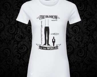 May include: A white short-sleeved t-shirt featuring a black graphic print. The design displays the text "FREAKSHOW PRESENTS: THE TALLEST IN THE WORLD" and "THE SMALLEST," with illustrations of a pair of long legs in suspenders and a small figure.