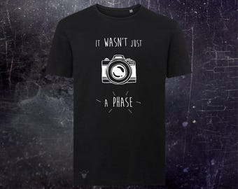 May include: A black short-sleeved t-shirt featuring a white graphic of a camera in the center. Above the camera, the text reads IT WASN'T JUST, and below the camera, the text reads A PHASE, with small radiating lines surrounding the lower text.