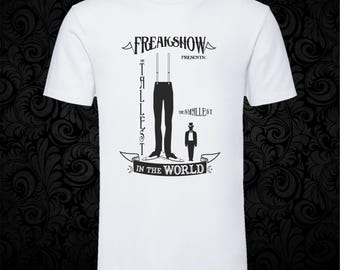 May include: A white short-sleeved t-shirt featuring a black graphic print that reads FREAKSHOW PRESENTS: THE TALLEST IN THE WORLD and THE SMALLEST. The design depicts a pair of long legs with suspenders and a small figure wearing a top hat.