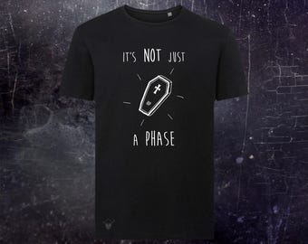 May include: A black short-sleeved t-shirt featuring a white graphic of a coffin with a cross on the lid. The text "IT'S NOT JUST A PHASE" is printed in white above and below the coffin illustration. The shirt has a round neckline and a small logo.