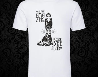 May include: A white short-sleeved t-shirt featuring a black graphic print of a figure in a long gown with ornate swirling patterns. The text reads THE AMAZING BEARDED LADY, accompanied by two crescent moon symbols and small star icons on the sides.
