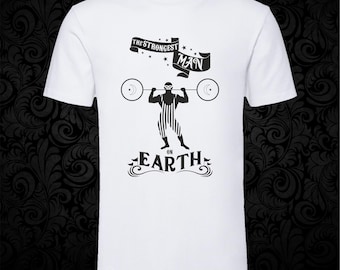May include: A white short-sleeved t-shirt featuring a black graphic of a masked weightlifter in a striped singlet lifting a barbell. Above the figure is a banner with the text THE STRONGEST MAN and below is the text ON EARTH with decorative swirls.