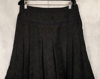 May include: A black knee-length skirt featuring a wide waistband with intricate tonal embroidery. The skirt has a pleated design throughout the lower section and a subtle, repeating floral jacquard pattern woven into the fabric of the garment.