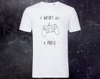 May include: A white short-sleeved t-shirt featuring a black line-art graphic of a video game controller centered on the chest. Above the controller, the text reads IT WASN'T JUST, and below the controller, the text reads A PHASE in black lettering.