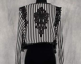 May include: A cropped jacket featuring a black and white vertical striped pattern. The garment is adorned with intricate black lace appliques along the shoulders, down the sides, and a large ornate lace design centered on the back panel.