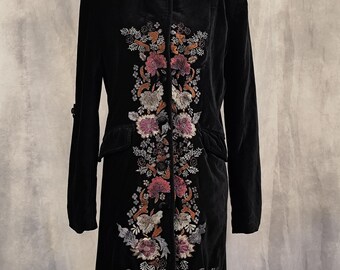 May include: A black velvet long coat featuring a high collar and long sleeves. The front of the coat is decorated with an intricate vertical floral embroidery pattern in shades of pink, orange, grey, and white, extending from the chest to the hem.