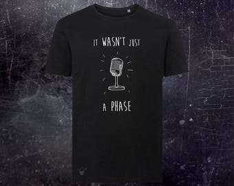 May include: A black short-sleeved t-shirt featuring a white line-art illustration of a vintage microphone in the center. Above the microphone is the text IT WASN'T JUST and below it is the text A PHASE. A small skull icon is visible at the bottom left.