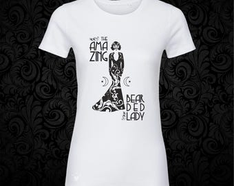May include: A white short-sleeved t-shirt featuring a black graphic print of a figure in a long gown with the text THE AMAZING BEARDED LADY. The design includes crescent moon and star symbols flanking the central figure on the front of the shirt.