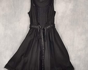 May include: A black sleeveless dress featuring a scoop neckline, ruffled shoulder straps, and a central vertical panel with lace-up detailing. The waist is cinched with a matching black belt featuring two metal rings, leading to a flared skirt.