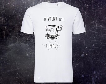 May include: A white short-sleeved t-shirt featuring a black line drawing of a top hat with internal gears and a pressure gauge. The text "IT WASN'T JUST A PHASE" is printed in black, with the words positioned above and below the hat graphic.