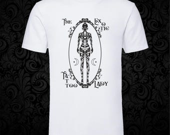 May include: A white short-sleeved t-shirt featuring a black graphic print of a person covered in intricate tattoos inside an ornate oval frame. The text "The Exotic Tattoo Lady" is printed in black around the central oval illustration.