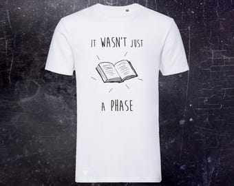 May include: A white short-sleeved t-shirt featuring a black line drawing of an open book in the center. Above the book, the text reads IT WASN'T JUST, and below the book, the text reads A PHASE. The design includes small radiating lines.