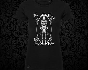 May include: A black short-sleeved t-shirt featuring a white graphic print of a figure covered in intricate tattoos inside an ornate oval frame. The text "The Exotic Tattoo Lady" is printed around the frame, accompanied by small crescent moon symbols.