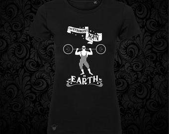 May include: A black short-sleeved t-shirt featuring a white graphic print of a masked weightlifter in a striped singlet lifting a barbell. Above the figure is a banner with the text THE STRONGEST MAN and below is the text ON EARTH.