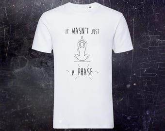 May include: A white short-sleeved t-shirt featuring a black line drawing of a person in a seated yoga pose. The text "IT WASN'T JUST A PHASE" is printed in black capital letters above and below the graphic, with small radiating lines.