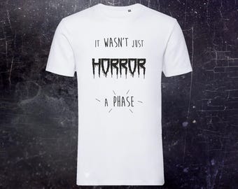 May include: A white short-sleeved t-shirt featuring black text that reads IT WASN'T JUST HORROR A PHASE. The word HORROR is printed in a stylized, dripping font, while the remaining text is in a simple, thin, sans-serif typeface.