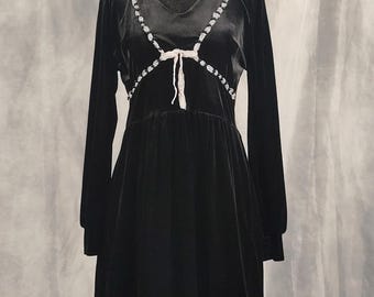 May include: A black long-sleeved velvet dress featuring a V-neckline, a gathered waist, and a ruffled hem. The dress is accented with white lace trim along the bodice and hem, featuring small white bows at the center chest and the skirt.