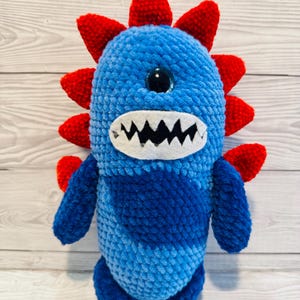 Op de afbeelding: Een pluche monster speelgoed gemaakt van blauw en donkerblauw garen. Het wezen heeft één groot zwart oog, een witte mond met gekartelde zwarte vilt tanden en een rij felrode stekels die langs de rug en de zijkanten van het lichaam lopen.