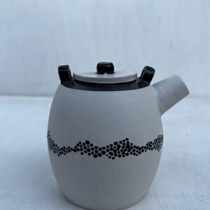 May include: A matte white ceramic teapot with a black lid and a black handle. The body features a horizontal band of small, black, hand-painted dots arranged in a wavy pattern. It has a short, wide spout on the right side and a flat lid.