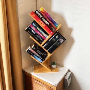 May include: A light brown wooden tree-shaped bookshelf with multiple angled shelves holding various books, including titles like Wayne Rooney Colour, Perfect Liars, The Teacher, and Harry Potter, with a small potted succulent at the base.