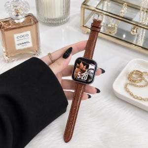 Op de afbeelding: Een smartwatch met een bruine, krokodil-getextureerde leren band en een zilverkleurige rechthoekige behuizing. Het digitale display toont de tijd 5:20 en een afbeelding van herfstbladeren. De band is voorzien van een zilverkleurige metalen gesp.