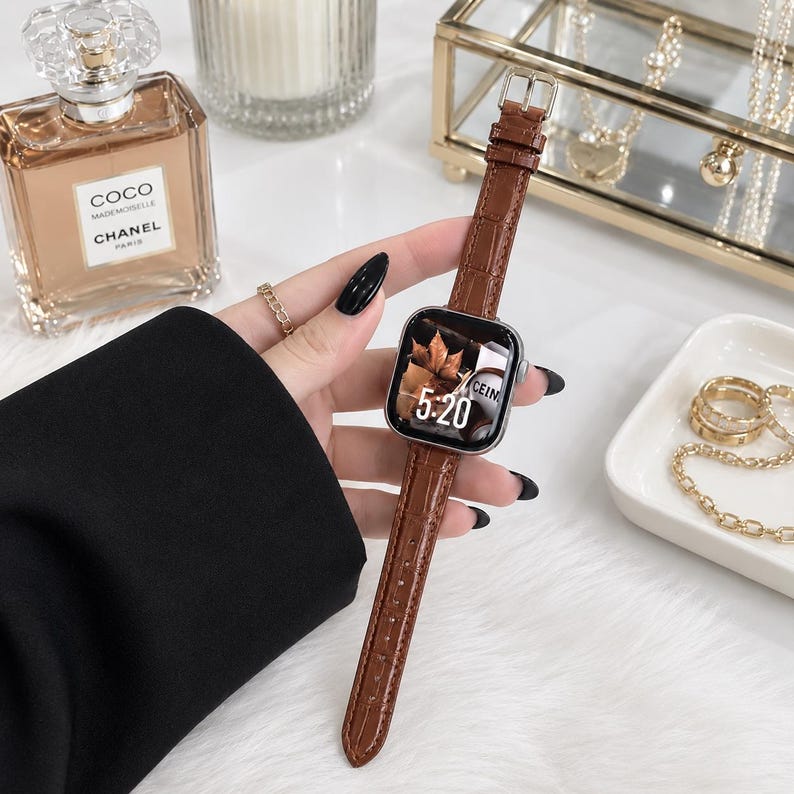 Op de afbeelding: Een smartwatch met een bruine, krokodil-getextureerde leren band en een zilverkleurige rechthoekige behuizing. Het digitale display toont de tijd 5:20 en een afbeelding van herfstbladeren. De band is voorzien van een zilverkleurige metalen gesp.