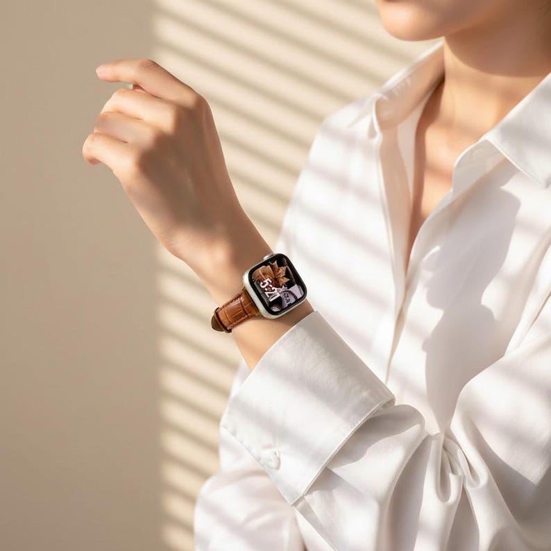 Op de afbeelding: Een zilveren smartwatch met een rechthoekige wijzerplaat en een bruine leren band met een textuur van krokodillenleer. Het display van het horloge toont een digitale klok die 5:20 aangeeft en een afbeelding van herfstbladeren.