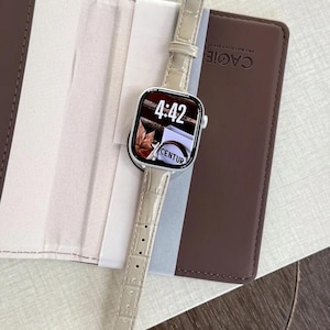 Op de afbeelding: Een smartwatch met een rechthoekige zilveren behuizing en een lichtbeige kunstlederen band met krokodillenmotief. Het digitale display toont de tijd 4:42 en de tekst CENTUR. Het horloge ligt op een bruine map met de merknaam CAGIE.