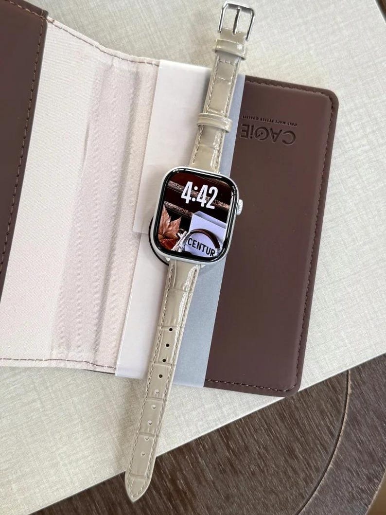 Op de afbeelding: Een smartwatch met een rechthoekige zilveren behuizing en een lichtbeige kunstlederen band met krokodillenmotief. Het digitale display toont de tijd 4:42 en de tekst CENTUR. Het horloge ligt op een bruine map met de merknaam CAGIE.