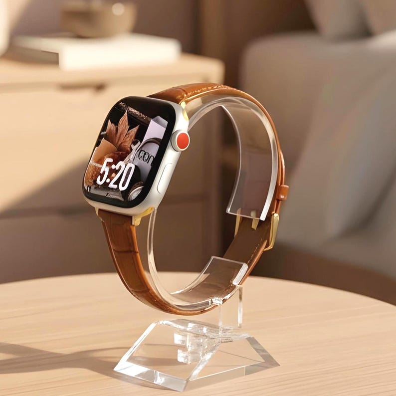 Op de afbeelding: Een zilveren smartwatch met een bruine leren horlogeband met krokodillenprint en een rode accentkleur op de zijknop. Het digitale scherm toont de tijd 5:20 en een afbeelding van herfstbladeren. Het horloge staat op een doorzichtige acrylaatstandaard.