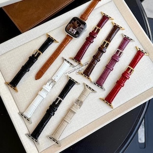 Op de afbeelding: Een collectie van negen horlogebandjes met een krokodil-reli&euml;f in diverse kleuren, waaronder zwart, wit, bruin, rood, bordeaux en beige. E&eacute;n bruin bandje is bevestigd aan een zilveren smartwatch die de tijd 5:24 op het scherm toont.