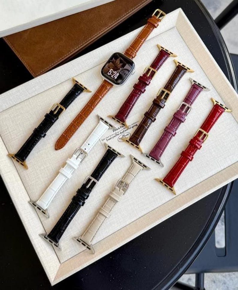 Op de afbeelding: Een collectie van negen horlogebandjes met een krokodil-reli&euml;f in diverse kleuren, waaronder zwart, wit, bruin, rood, bordeaux en beige. E&eacute;n bruin bandje is bevestigd aan een zilveren smartwatch die de tijd 5:24 op het scherm toont.