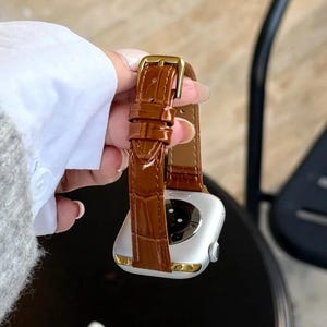Op de afbeelding: Een zilverkleurige smartwatch met een glanzende bruine leren horlogeband met een krokodil-reli&euml;f en een goudkleurige metalen gesp. De horlogekast heeft een gladde, metalen afwerking met een cirkelvormige sensor aan de achterzijde.