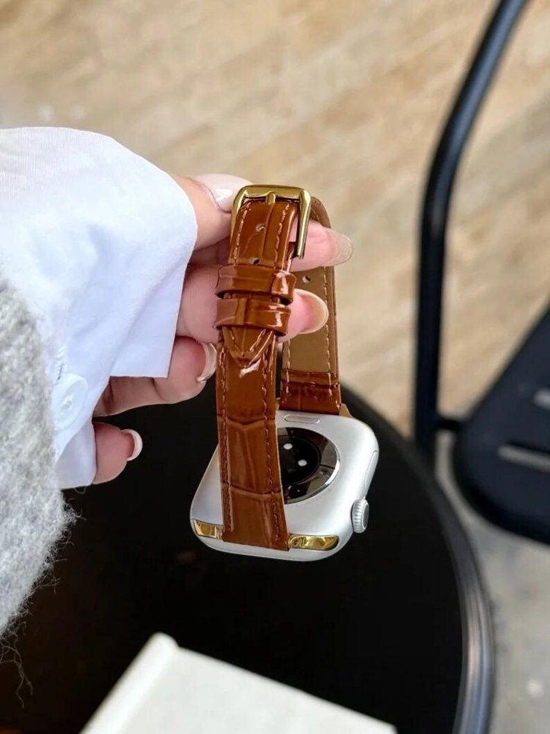 Op de afbeelding: Een zilverkleurige smartwatch met een glanzende bruine leren horlogeband met een krokodil-reli&euml;f en een goudkleurige metalen gesp. De horlogekast heeft een gladde, metalen afwerking met een cirkelvormige sensor aan de achterzijde.
