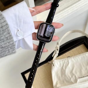 Op de afbeelding: Een smartwatch met een zilverkleurige rechthoekige behuizing en een glanzende zwarte kunstlederen band met krokodillenmotief en een goudkleurige gesp. De digitale wijzerplaat toont de tijd 4:58 met een monochrome achtergrondafbeelding.
