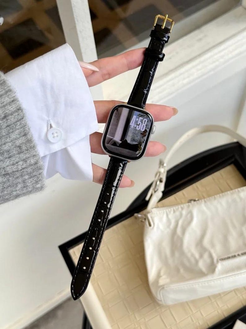Op de afbeelding: Een smartwatch met een zilverkleurige rechthoekige behuizing en een glanzende zwarte kunstlederen band met krokodillenmotief en een goudkleurige gesp. De digitale wijzerplaat toont de tijd 4:58 met een monochrome achtergrondafbeelding.
