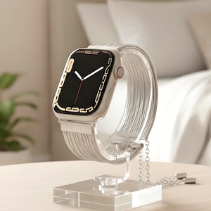 Puede incluir: Un reloj inteligente plateado con pantalla digital negra, manecillas blancas y segundero rojo. El reloj tiene una correa metálica de múltiples hilos finos y está montado sobre un soporte de exhibición de acrílico transparente con base cuadrada.
