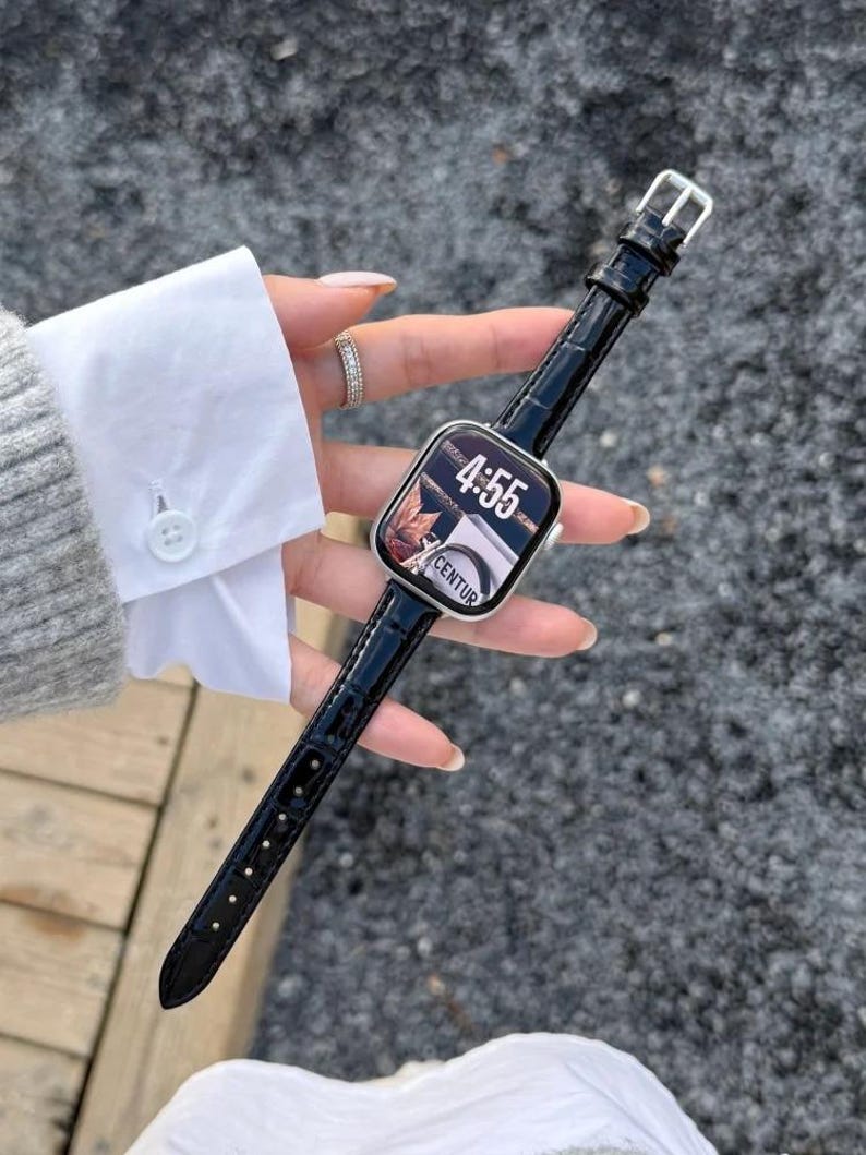 Op de afbeelding: Een smartwatch met een rechthoekige zilveren behuizing en een zwarte lederen horlogeband met krokodillenprint. Het digitale display toont de tijd 4:55 en de tekst CENTUR. Het horloge heeft een zilveren gesp en een gepolijste afwerking.