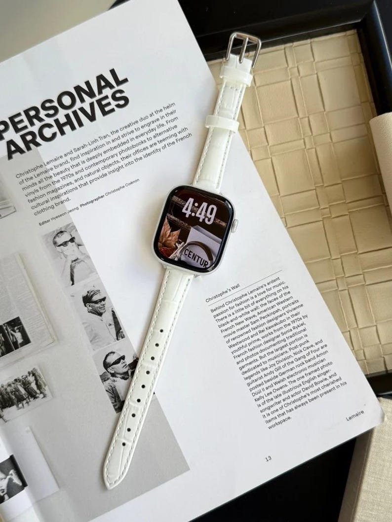 Op de afbeelding: Een smartwatch met een witte, gestructureerde leren band en een zilverkleurige behuizing. Het digitale display toont de tijd 4:49 en een afbeelding met de tekst CENTUR. Het horloge ligt verticaal op een open tijdschriftpagina met tekst.
